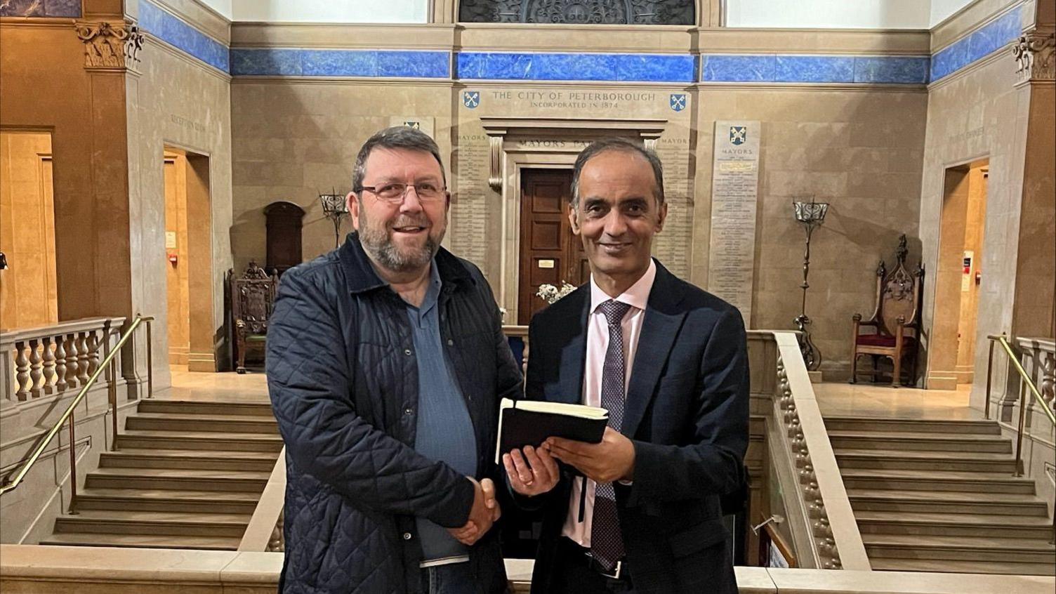 Mohammed Farooq and Chris Harper jointly hold a black book, while they stand in front of a grand staircase. The words "The City of Peterborough" are above a door behind them. Farooq is dressed in a dark suit, and Harper is wearing a blue polo shirt and navy jacket.    