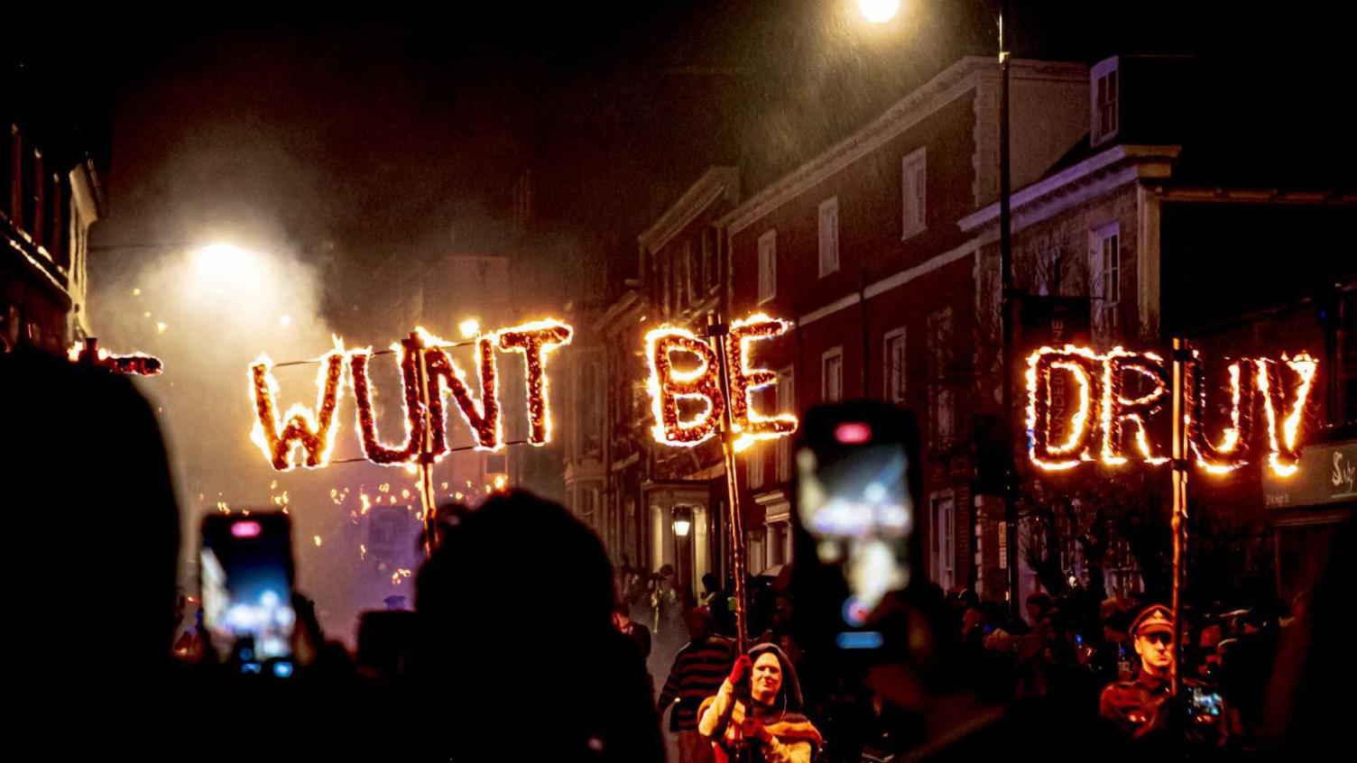 Words made up of letters that have been set on fire. People are carrying the words "wunt" "be" and "druv". At the forefront of the image are lots of silhouettes of people watching and filming on their phones. 