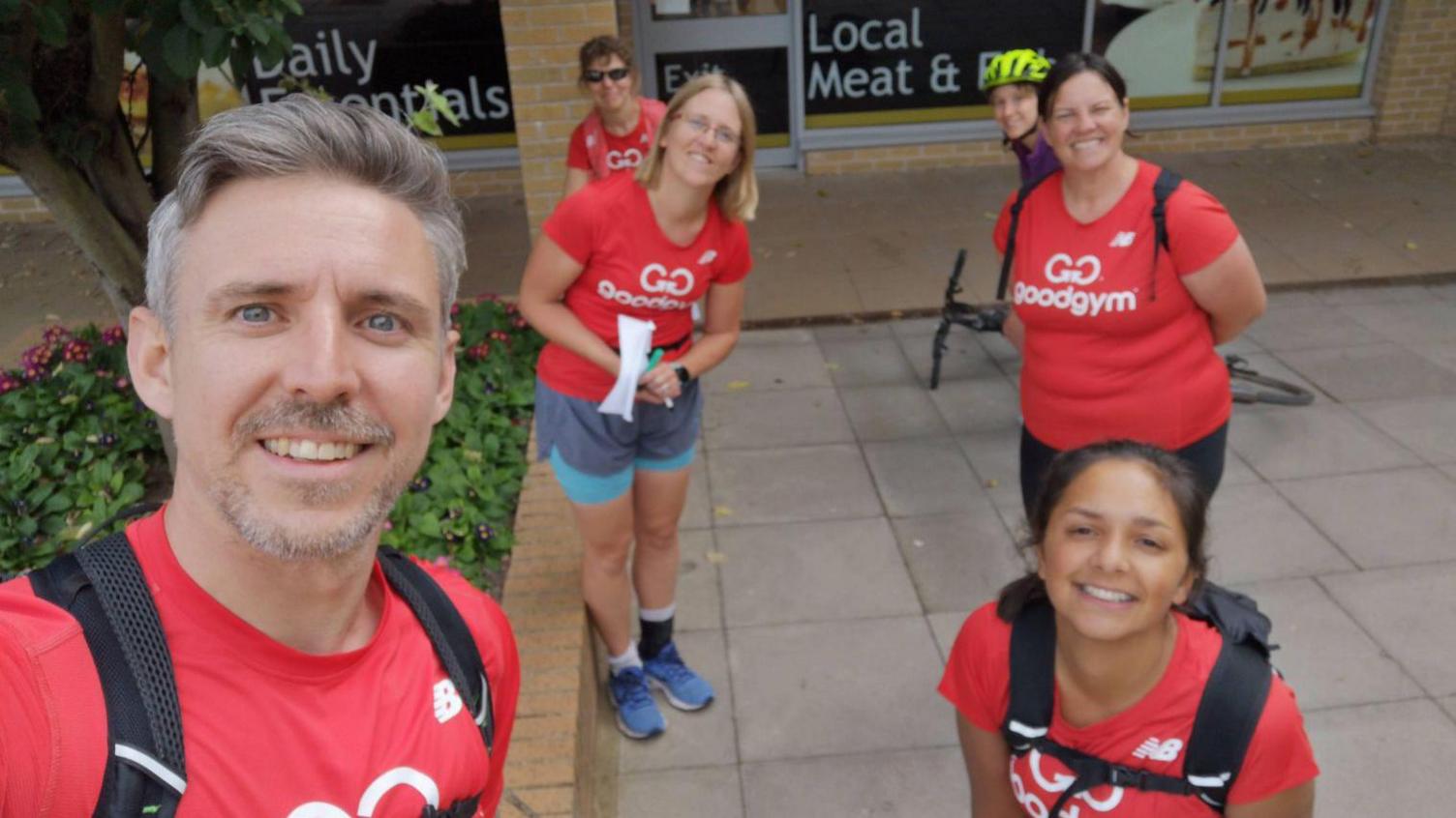 A man and five women in red T shirts that read 'GoodGym' in white wearing fitness clothing standing on a street.