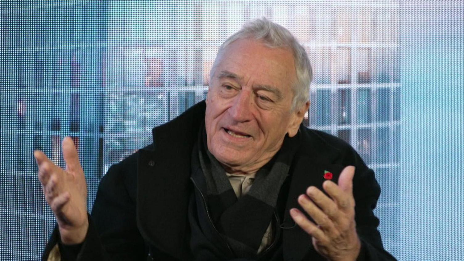 Robert De Niro, sitting in front of a blue background, wears a black coat and a dark scarf. He has short grey hair and is gesturing with both hands out in front of him.