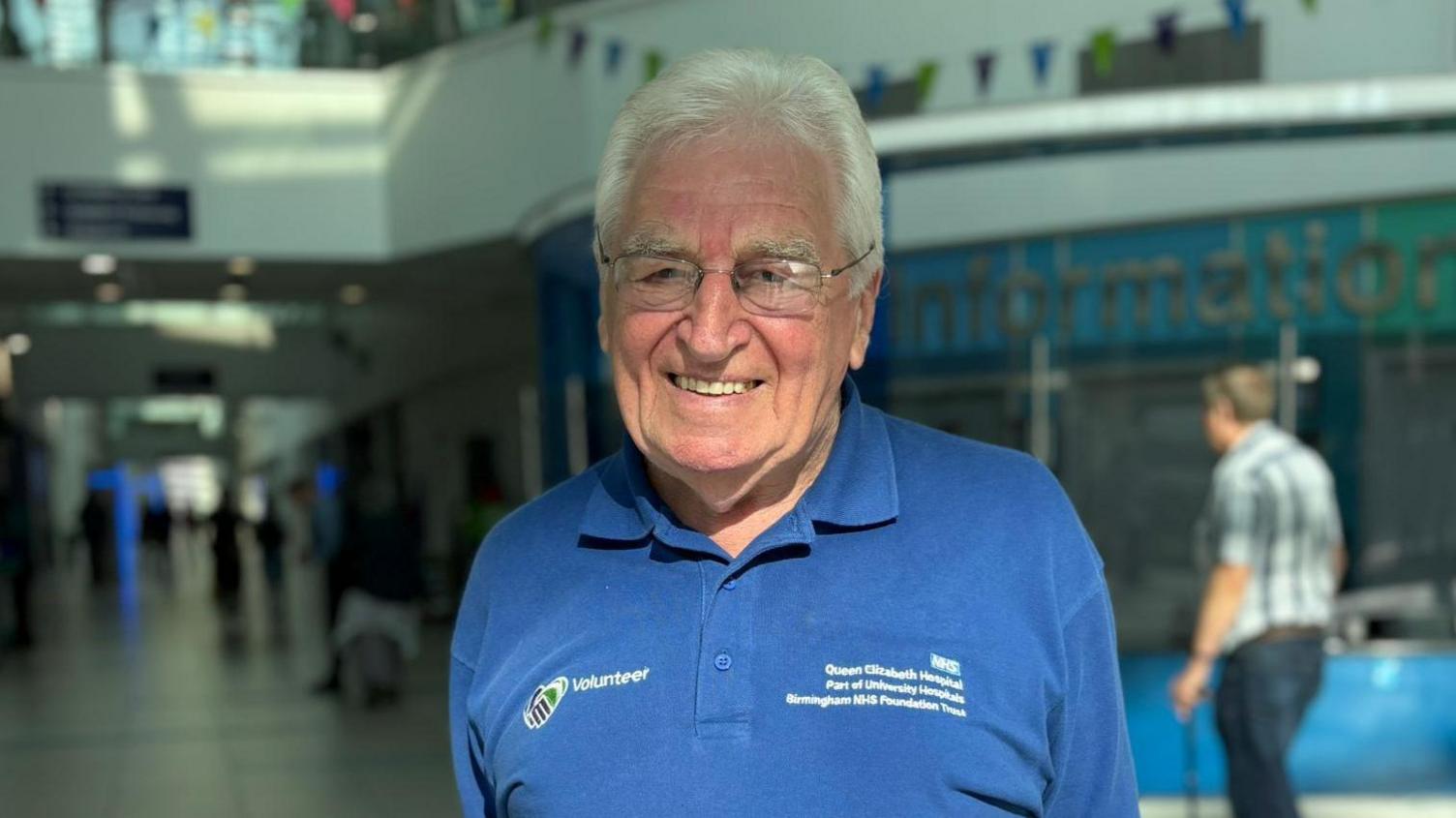 A man is a dark blue t-shirt with a "volunteer" sign on it. He is wearing glasses and smiling and the hospital is in the background. Another man is out of focus in the right of the picture.