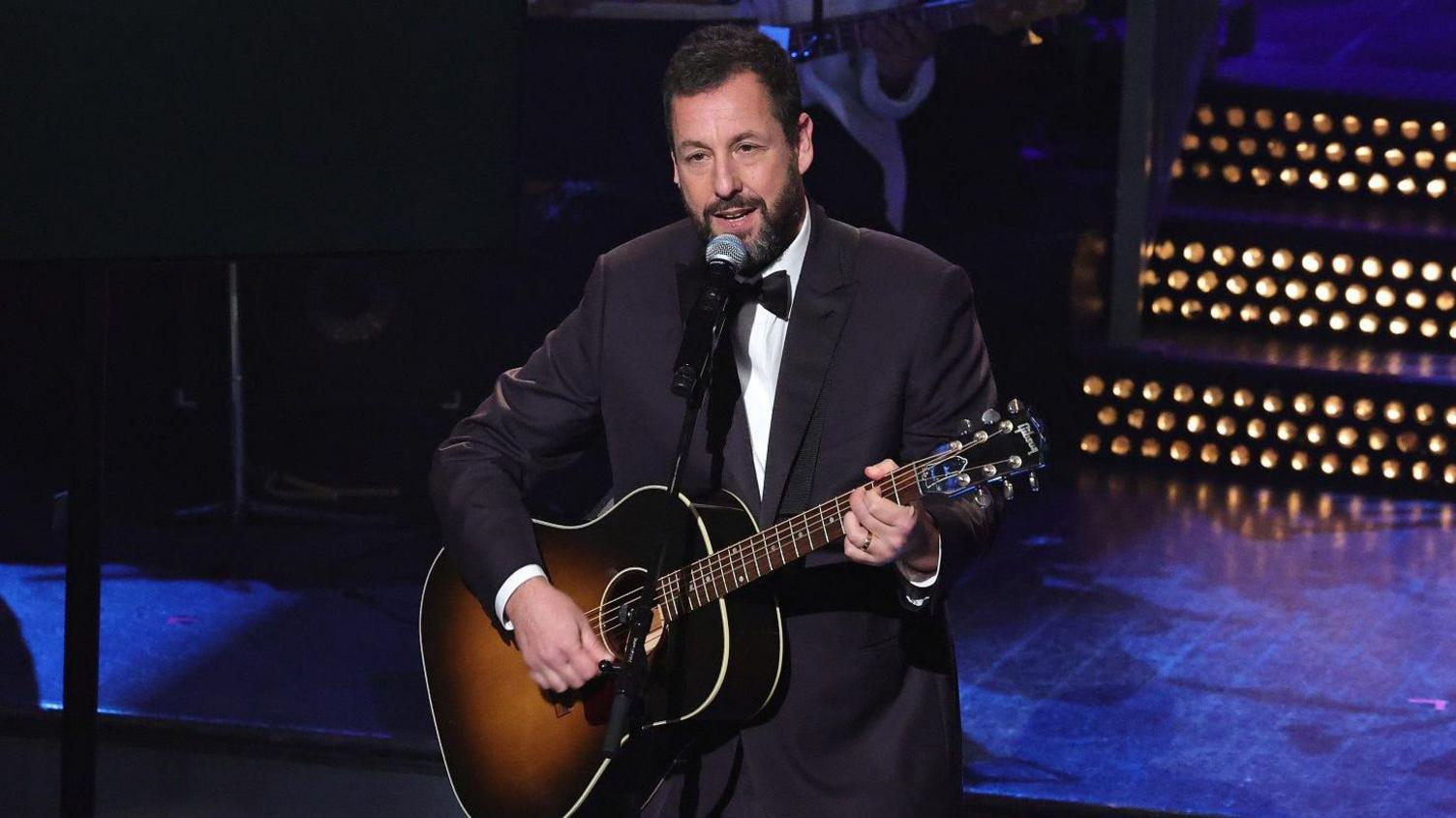 Adam Sandler wearing a tuxedo and playing an acoustic guitar