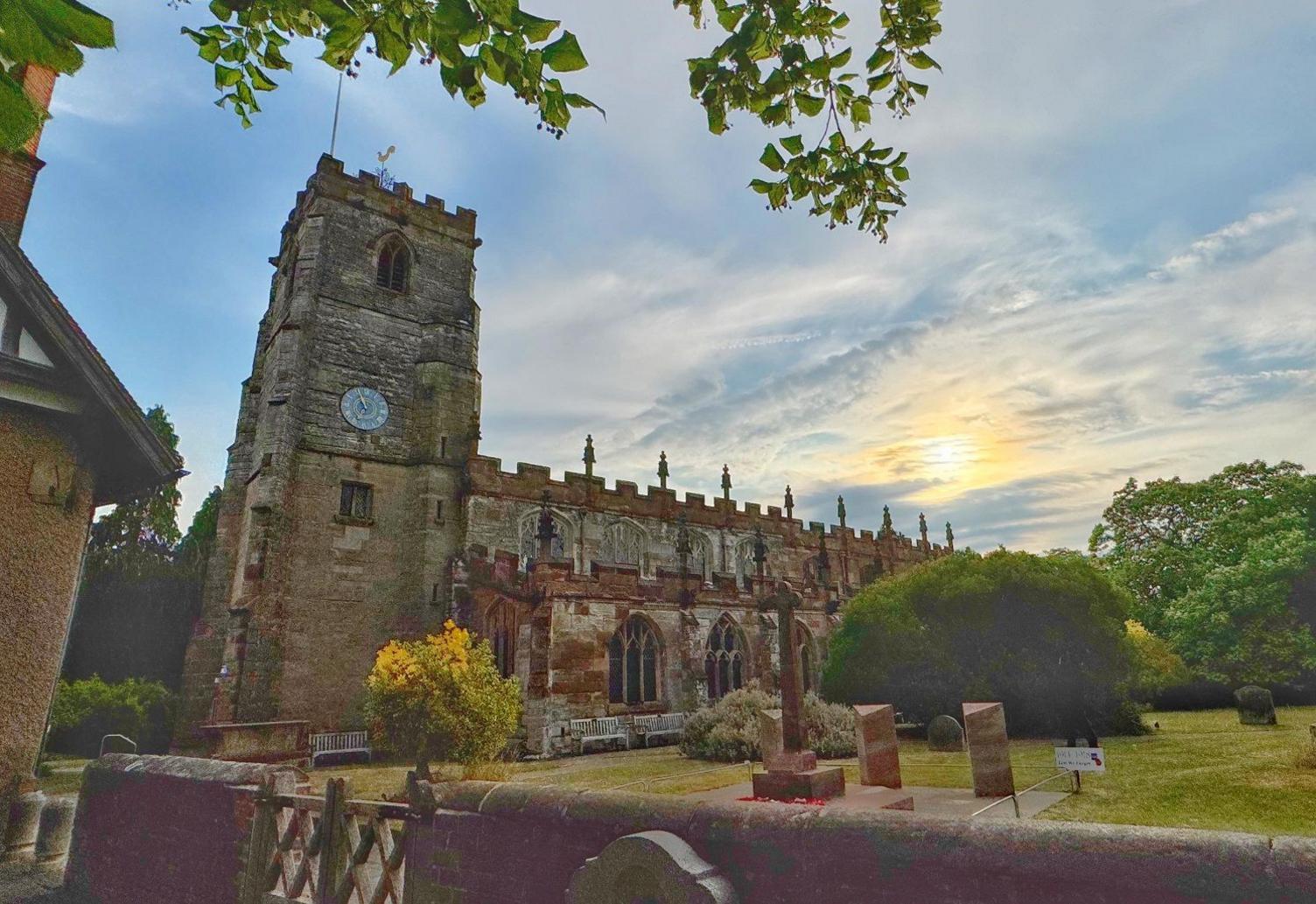 A church with a stone tower sits among a green area with a stone wall in front.