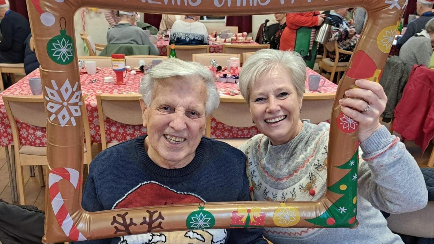 An elderly man with white hair and a broad smile looks at the camera alongside his daughter, who also has white hair. Both are wearing Christmas jumpers and are holding up an inflatable festive photo frame. 