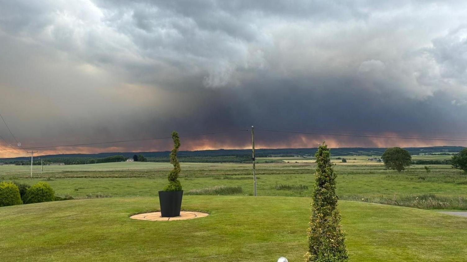 On the horizon, thick grey smoke rises from wildfires into a grey cloudy sky. The picture is taken over a lawn with carefully trimmed bushes.
