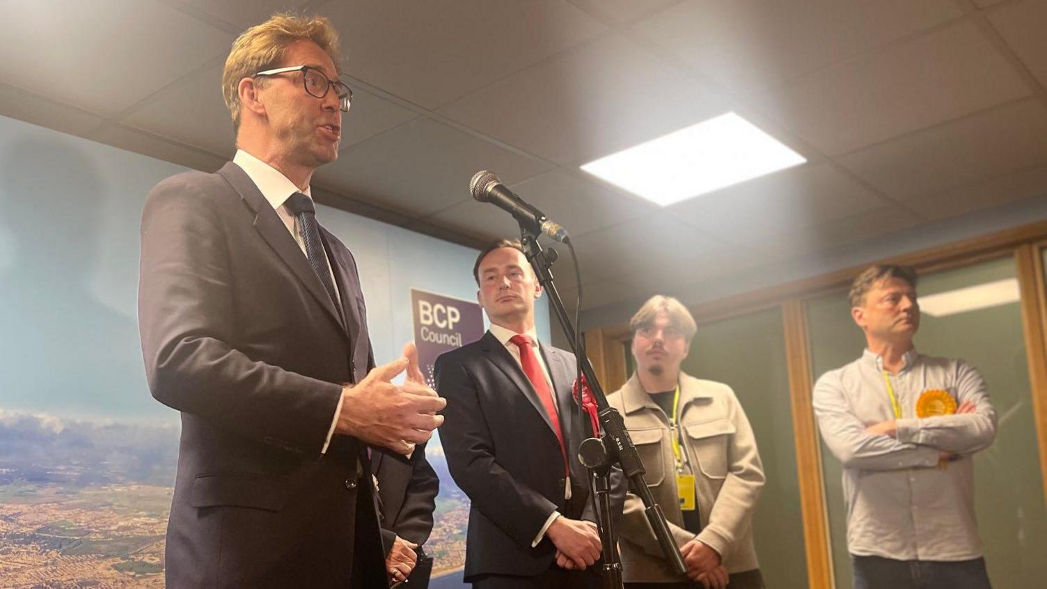 Tobias Ellwood speaking into a microphone. To his right are the other election candidates, including Tom Hayes