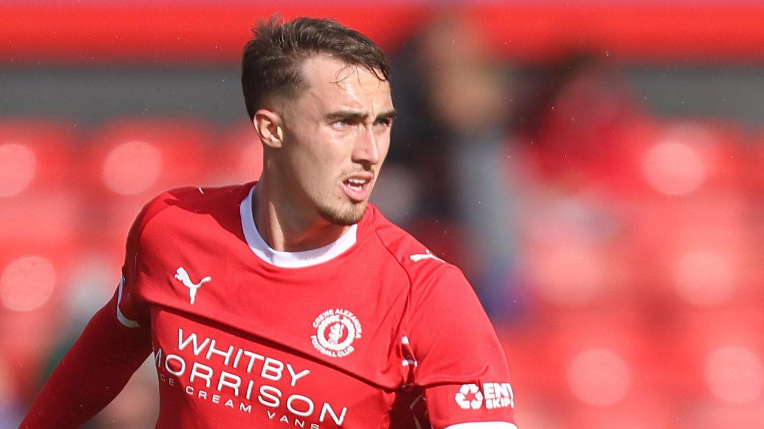 Lewis Billington in action for Crewe. He is wearing a red home shirt and is looking to his left preparing to pass the ball