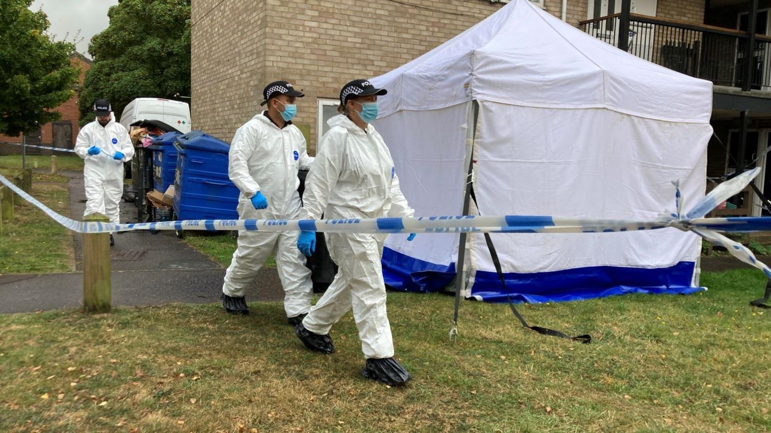 Three forensic investigators wearing white jumpsuits walking near a forensic tent outside a block of flats