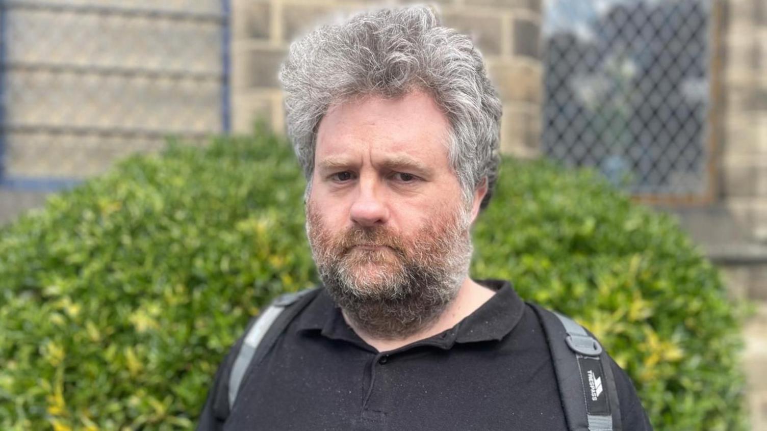 A man is wearing a black polo shirt on and a Trespass backpack with grey straps. He has curly grey hair and a brown and grey beard. He is stood in front of a big green bush outside a brick building.
