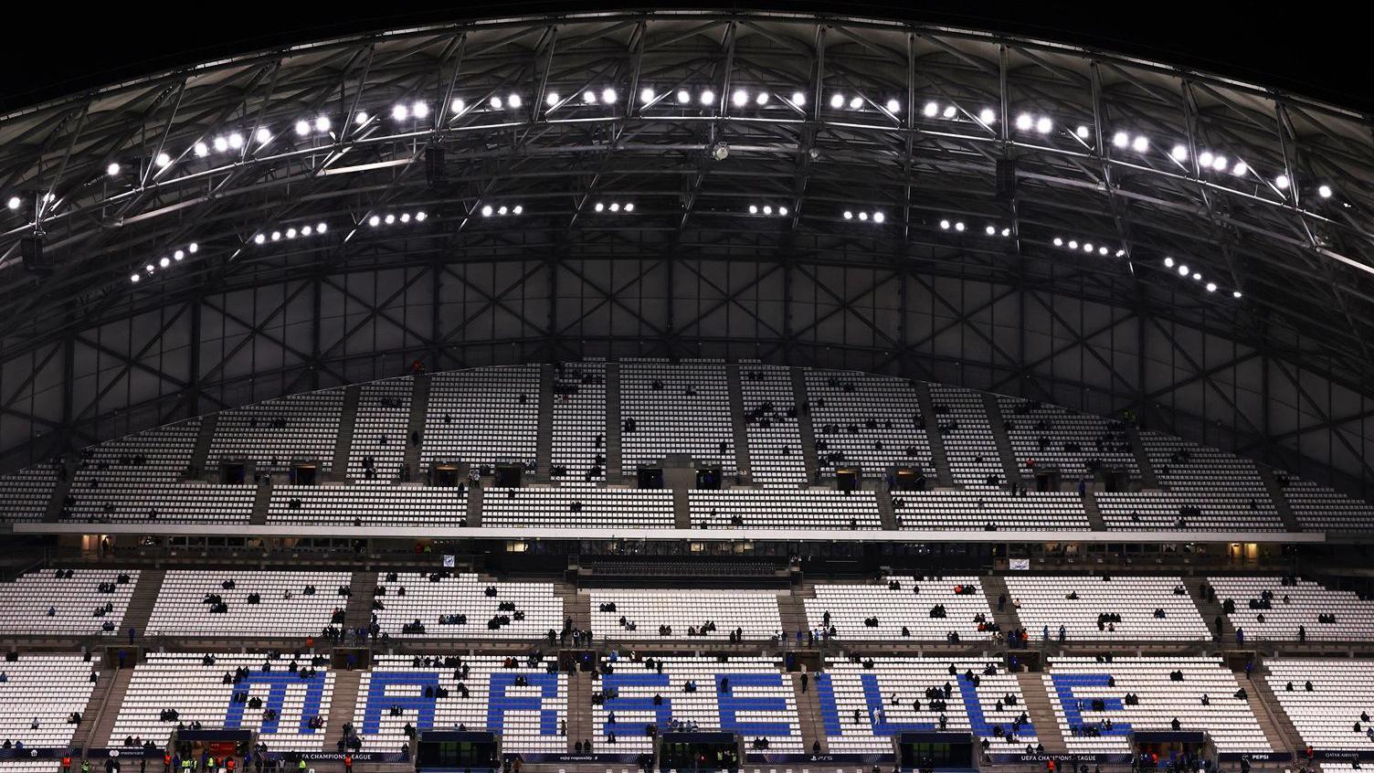 Inside the Stade Vélodrome showing one of the stands. The stand has a curved roof and is filled with white chairs. Towards the bottom Marseille is spelt out in blue chairs. Groups of people are sat in their seats but it is largely empty. It is dark and the stadium lights are on.