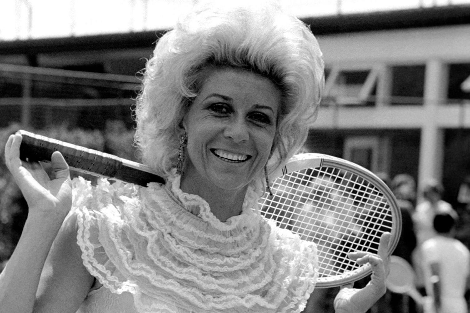 A black and white photo of Lea Pericoli holding a tennis racket behind her shoulders. She is wearing bold earrings and a very frilly tennis top.