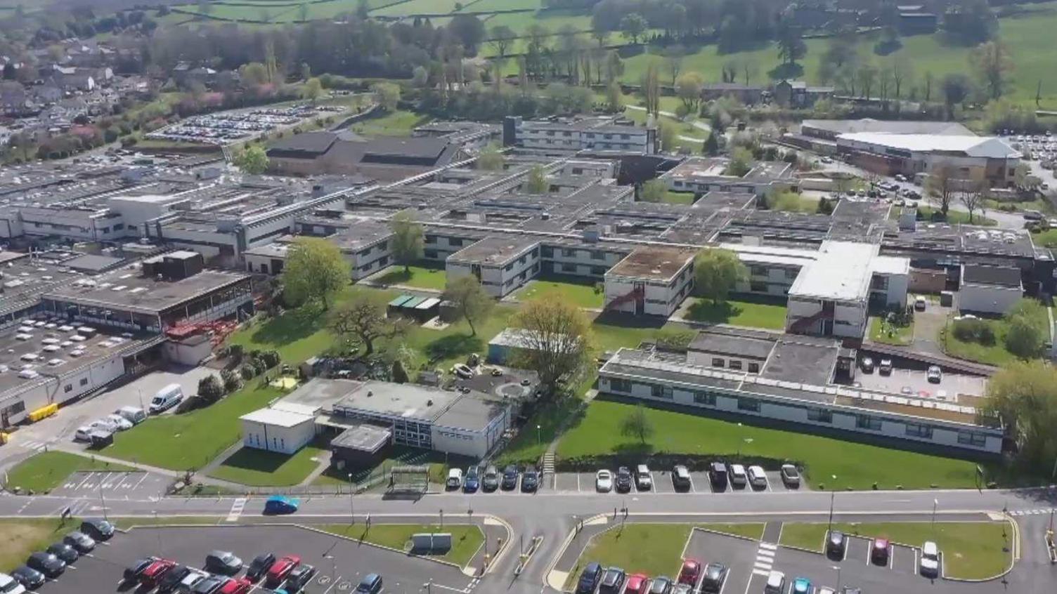 An aerial view of Airedale Hospital: car parks and green spaces can be seen in the foreground with an array of grey and white two-storey buildings behind. 