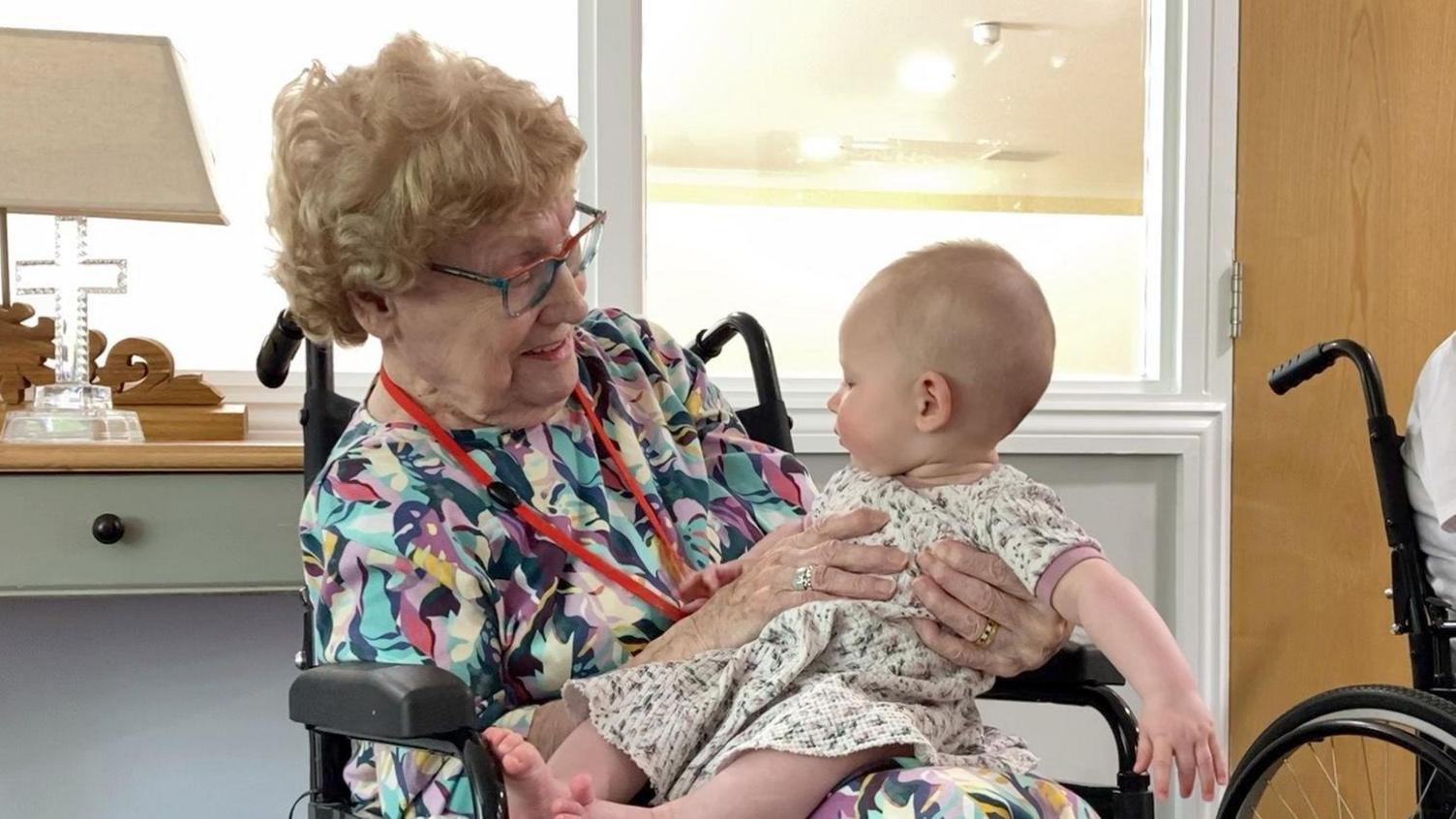 An older lady in a wheelchair with brightly coloured blouse holds a six-month-old baby who is playing with her lanyard. She sits in a wheelchair as we can see the black handles behind her head and the armrests to the sides.