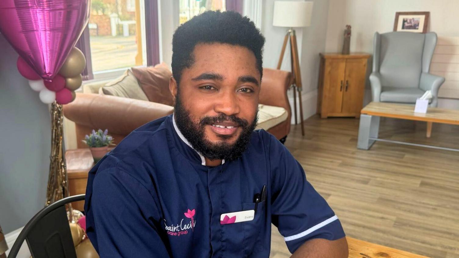 A man in a navy blue nurse uniform sits and smiles in a nursing home lounge area.