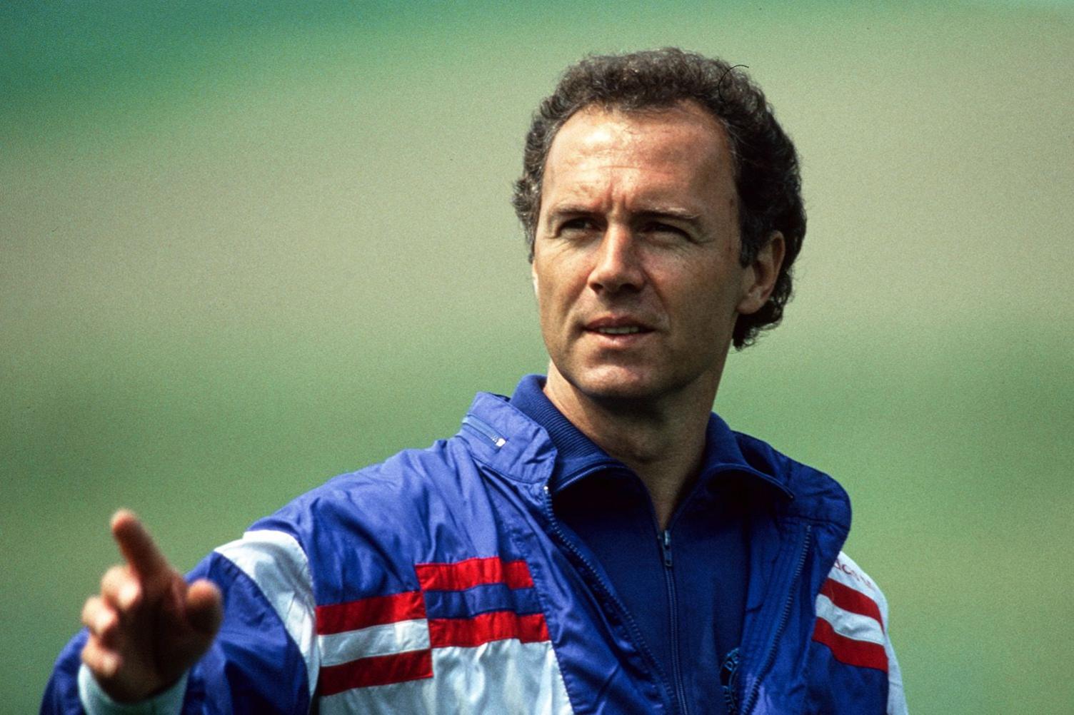 A picture of Franz Beckenbauer taken during his days as a coach - he has dark curly hair, is clean-shaven and is wearing a blue and white waterproof jacket with red horizontal stripes. He appears to be outdoors against a blurry green background and is pointing at something with his right hand. 