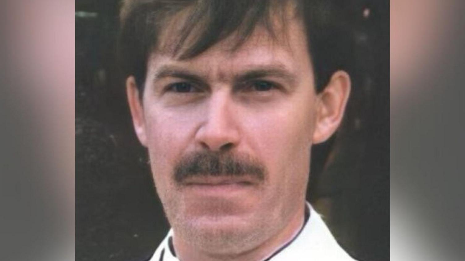 A man with a long face, brown eyes, a brown moustache, and straight brown hair over his forehead.