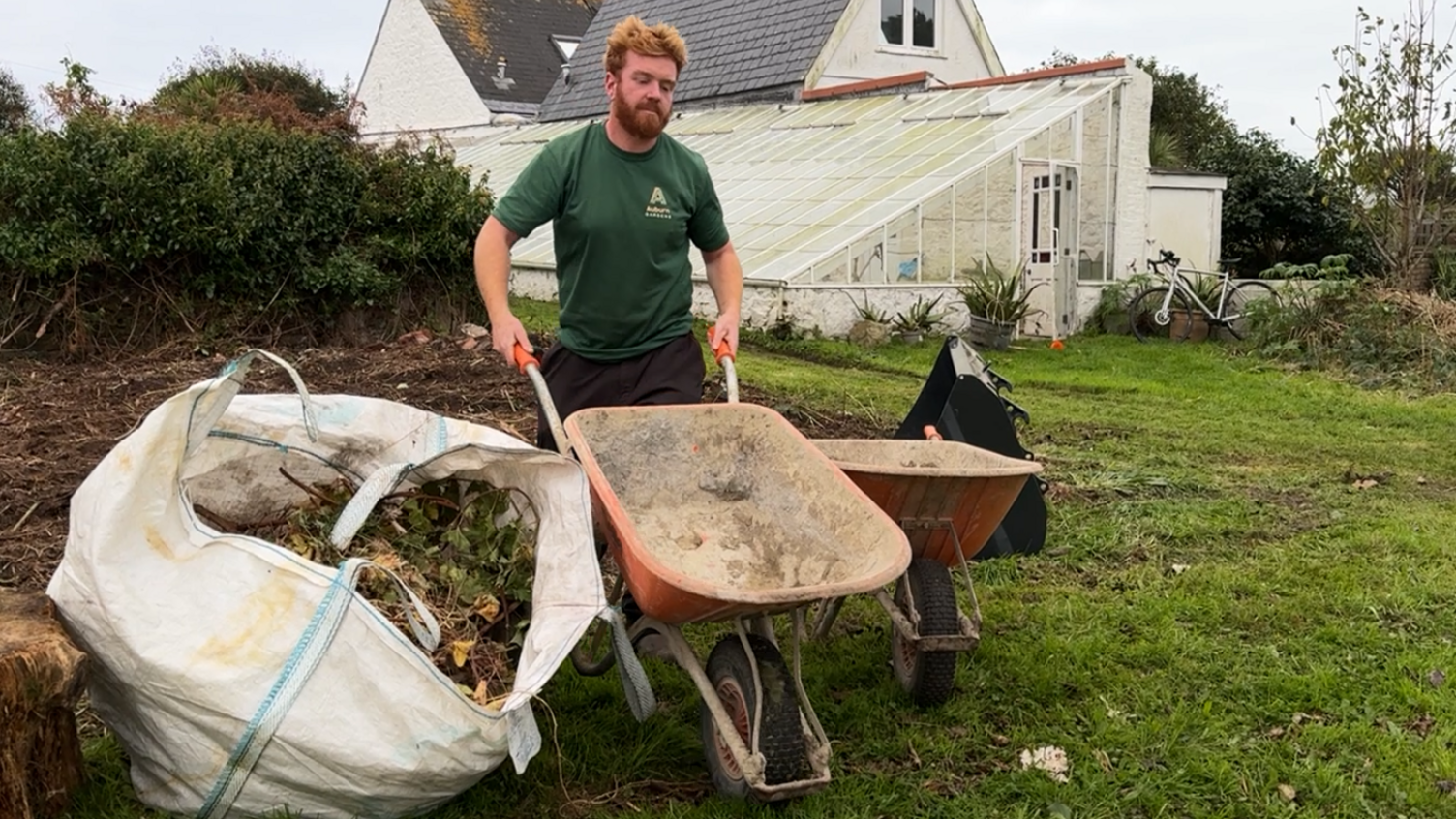 Jack Etheridge, wearing a green T-shirt, pushing a wheelbarrow beside a bag of green waste