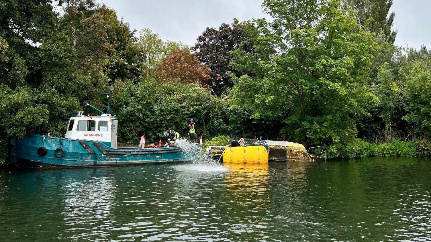 A view of a small boat next to the riverbank which has a large yellow flotation bag next to it. A water pump on another boat along side it is spraying out water in to the ricer.