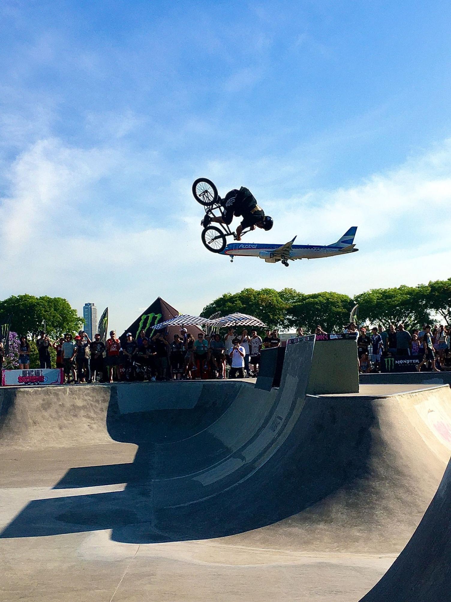 A BMX rider performs a backflip in a skate rink to a crowd. In the background a plane is landing. It makes it look like the bike is balancing on its front wheel on the nose of the plane.