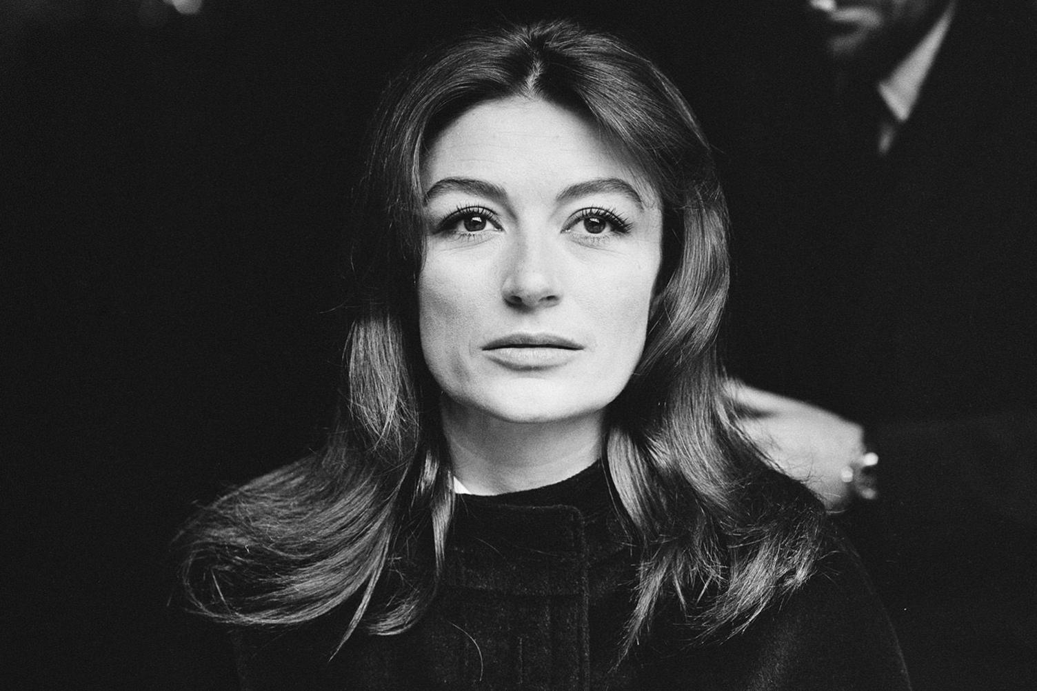 1960s news photo of Anouk Aimée, when she would have been in her early 30s. She is captured close-up and is staring ahead in a very confident manner. She has long dark hair and is wearing something black, which is indistinguishable from the very dark background of the photo.