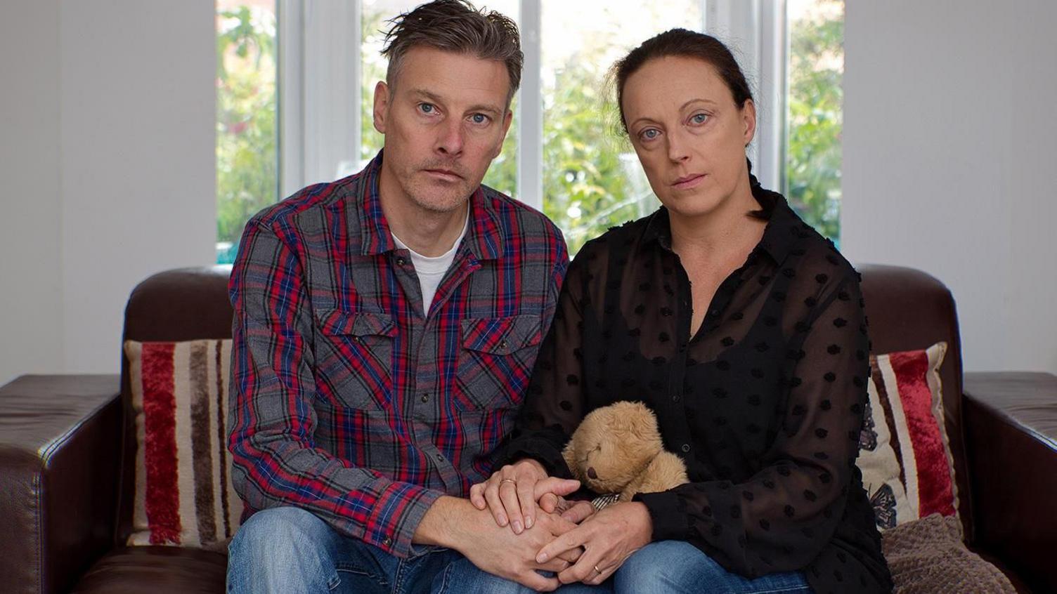 A man with short, styled brown hair and wearing a red, blue and grey checked shirt sits on a sofa next to his partner, a woman with tied back brown hair and wearing a sheer black top. They are holding hands and she has a teddy bear on her lap.