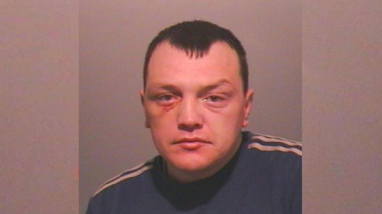 A mugshot of Gary Hughes. He has close-cropped dark hair, receding either side of a brushed forward centre point. He is wearing a blue sweatshirt with white stripes along the top of the shoulders and has a bruise under his right eye.