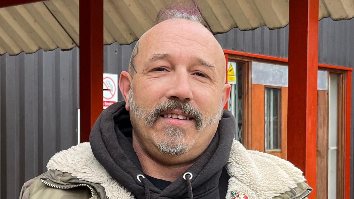 Carl Higgs is stood outside and is looking directly at the camera while smiling. He is predominantly bald with a patch of grey and purple hair in the centre of his head. He has a tattoo on the left side of his head and a grey facial hair. He wears a dark grey hoodie with a camo-style coat and poppy pin attached. 