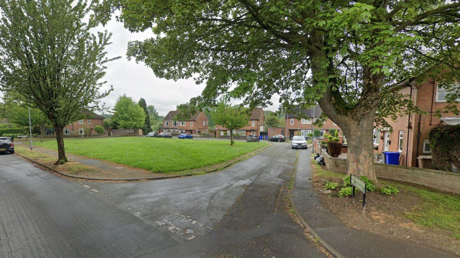 A Google street view image showing the entrance to a residential street. The road curves around to the left with houses on the right and a small, grassy area to the left. There are trees either side of the street.