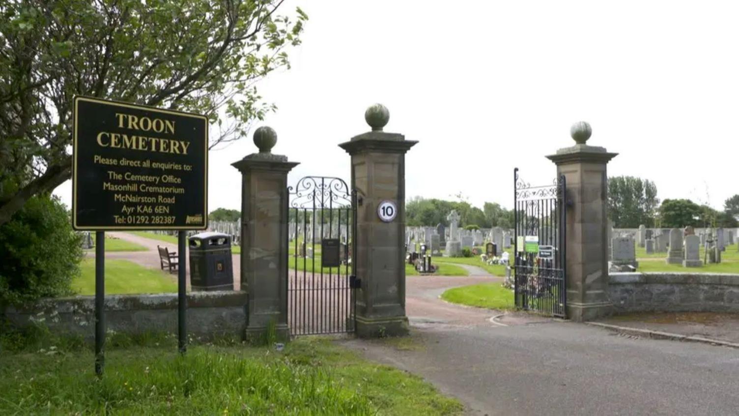 Image of the gates to Troon cemetery - a black sign with gold lettering stands on the left in front of the stone pillars of the gates and the iron gates themselves which are open. The graveyard is seen on the other side.