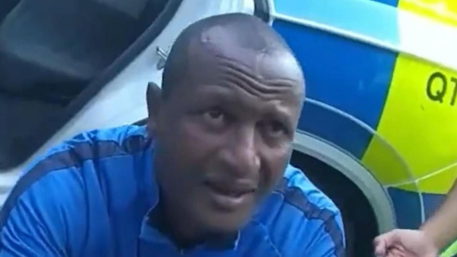 Hadush Kebatu has short black hair and is wearing a blue sports top. He is crouched in front of a police car and has his eyebrows raised.