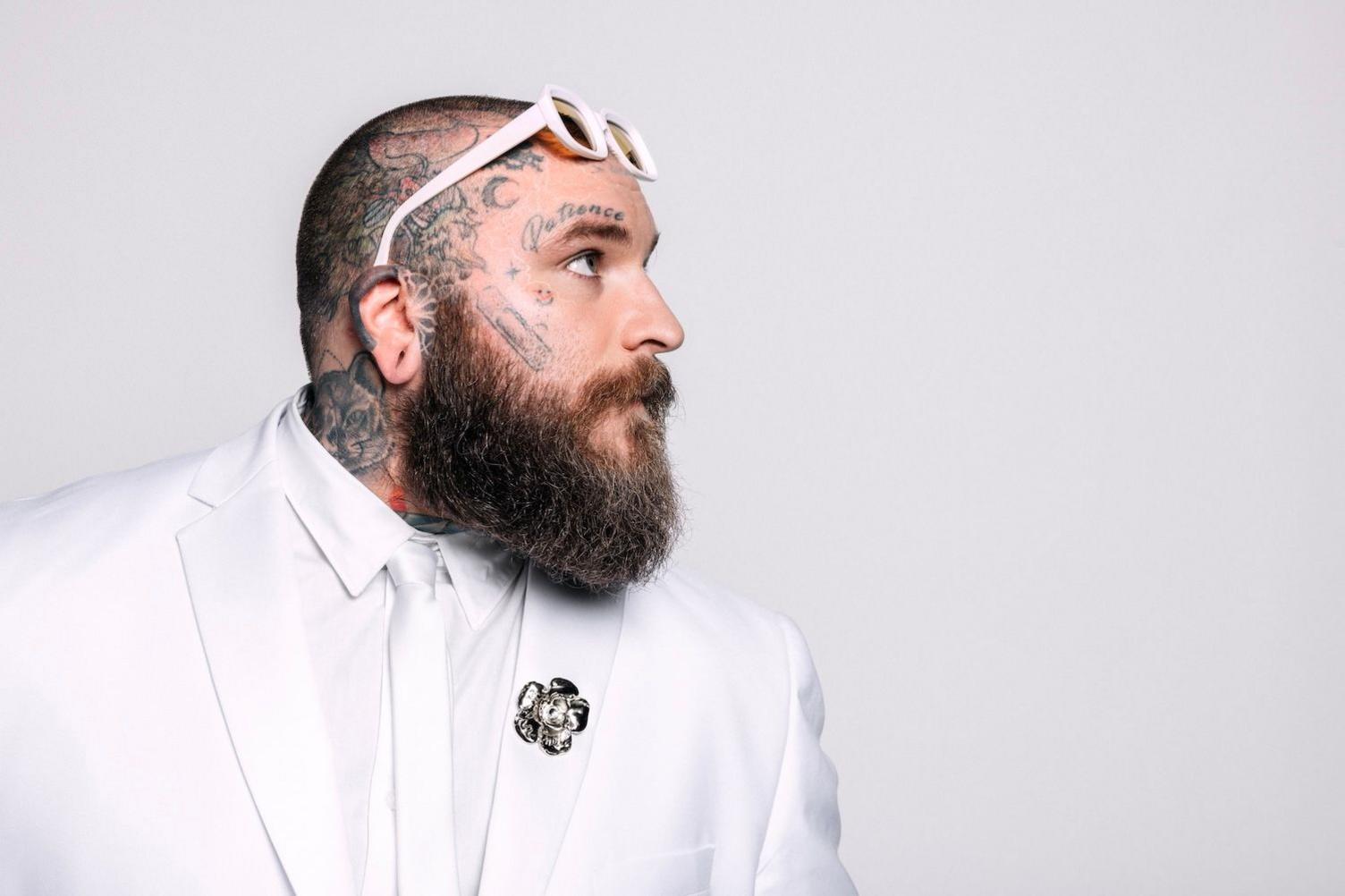 Teddy Swims, dressed in a white tux, poses sideways-on, with a pair of white glasses perched on his tattooed head