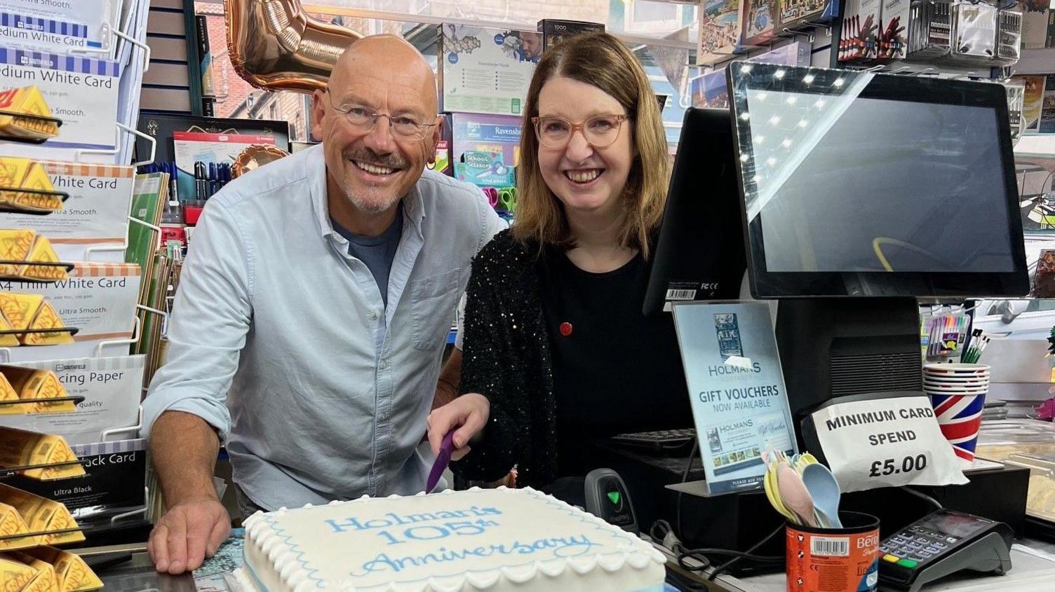 Inside a shop, two people stand behind a counter with a large rectangular cake decorated with white icing and a blue ribbon, featuring the words ‘Happy Anniversary.’ The counter is covered with illustrated maps and designs, and nearby are gift cards, stationery items, and a display of snacks.