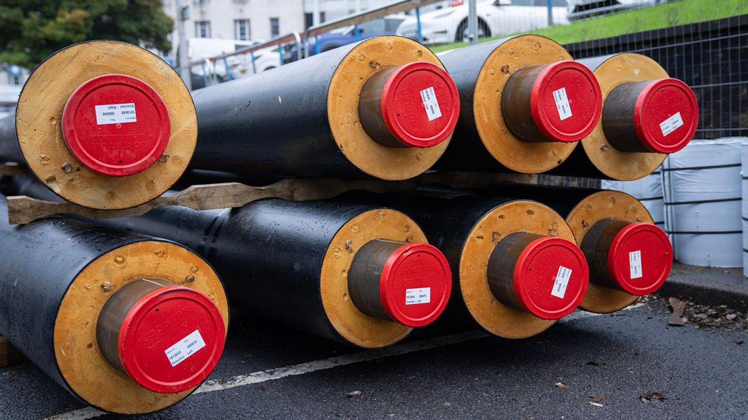 Eight long circular pipes piled on top of each other on a road. They are grey in colour with red on the end.