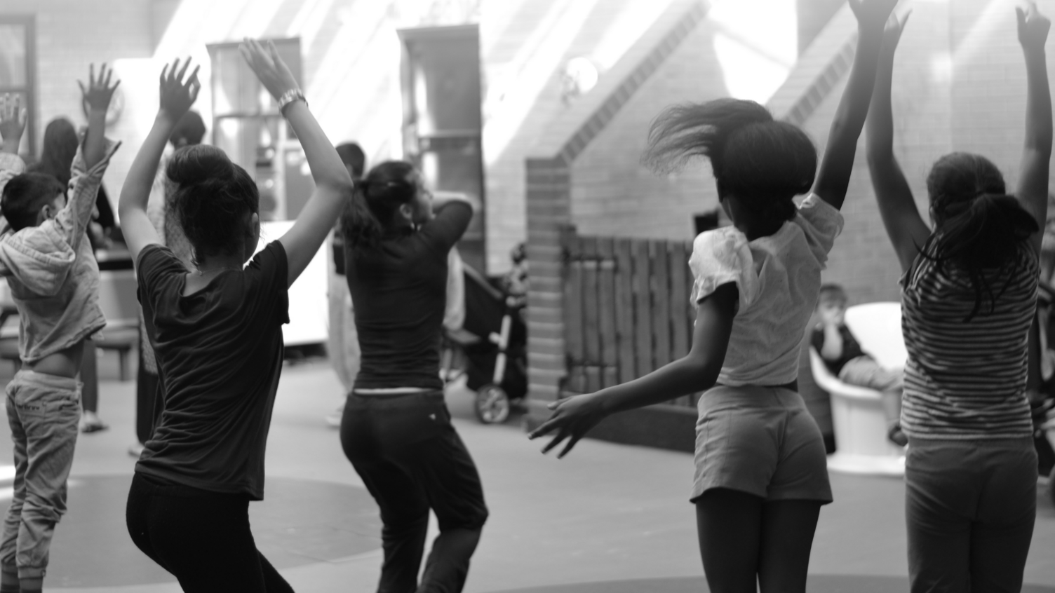 Several women and girls have their hands in the air with their backs to the camera. They look to have been dancing as they stand in an open area with a brick staircase at the far end of the room. The women wears dark trousers and tops, one of the girls has shorts and a white top, they all have dark hair.