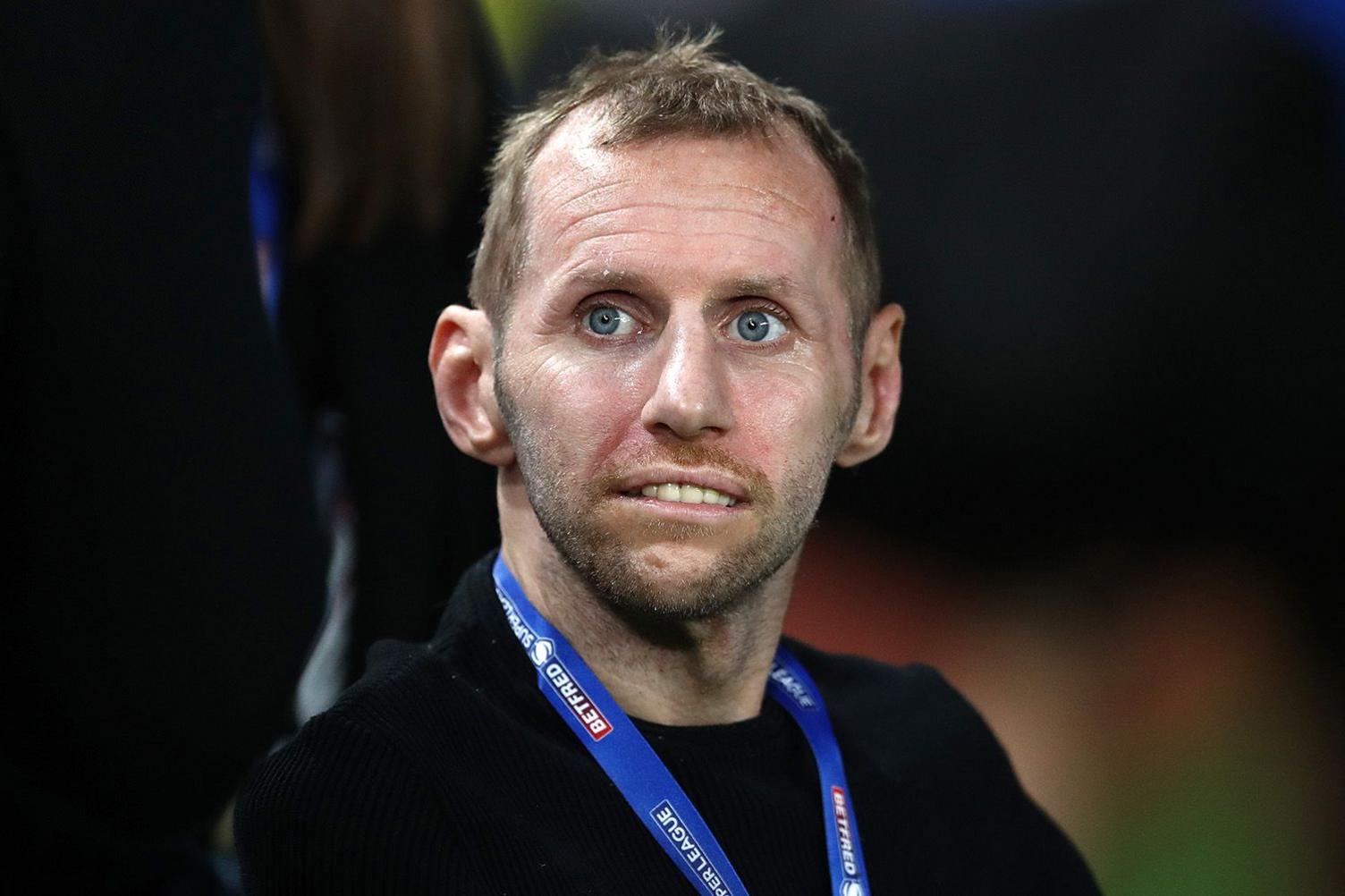 Rob Burrow pictured during the last few years of his life when he was suffering from MND - he has receding straw-coloured hair and he is looking beyond the camera, biting his lip. He seems to be in a wheelchair being pushed by an indistinct figure, whose lower half is visible in the photo. Rob Burrows wears a black sweater, with a blue lanyard around his neck.