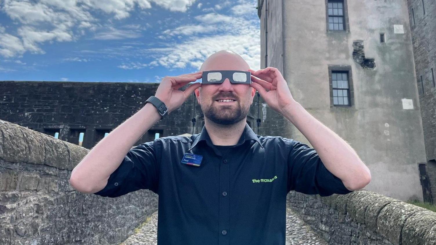 A man standing in front of Broughty Castle wearing eclipse glasses. He has on a dark blue shirt.