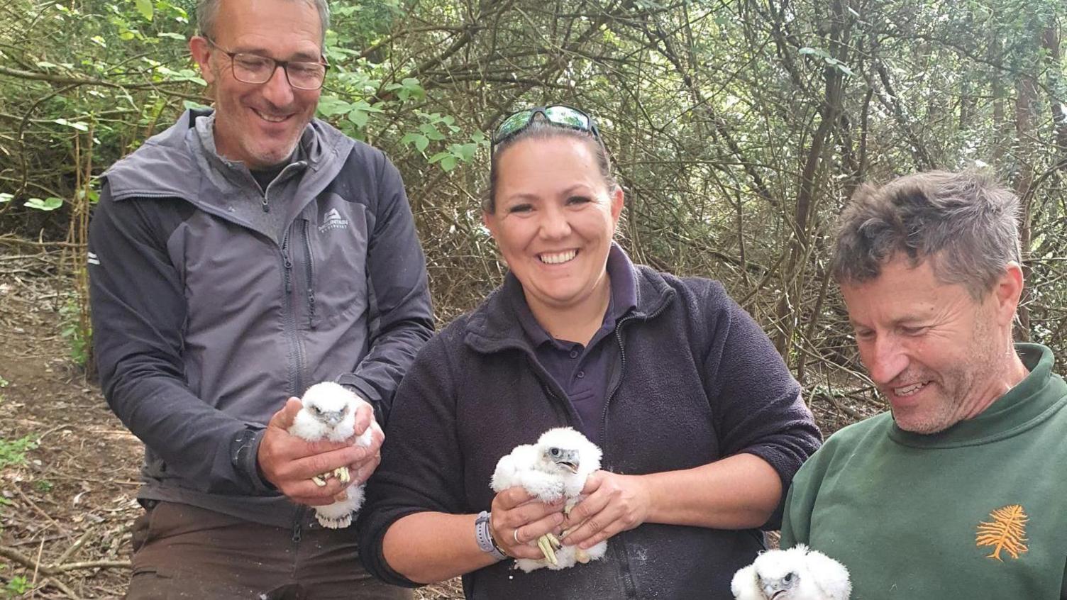 Two men wither side of a woman, stood in a forest. The three people are each holding a white falcon chick with a grey beak using two hands.