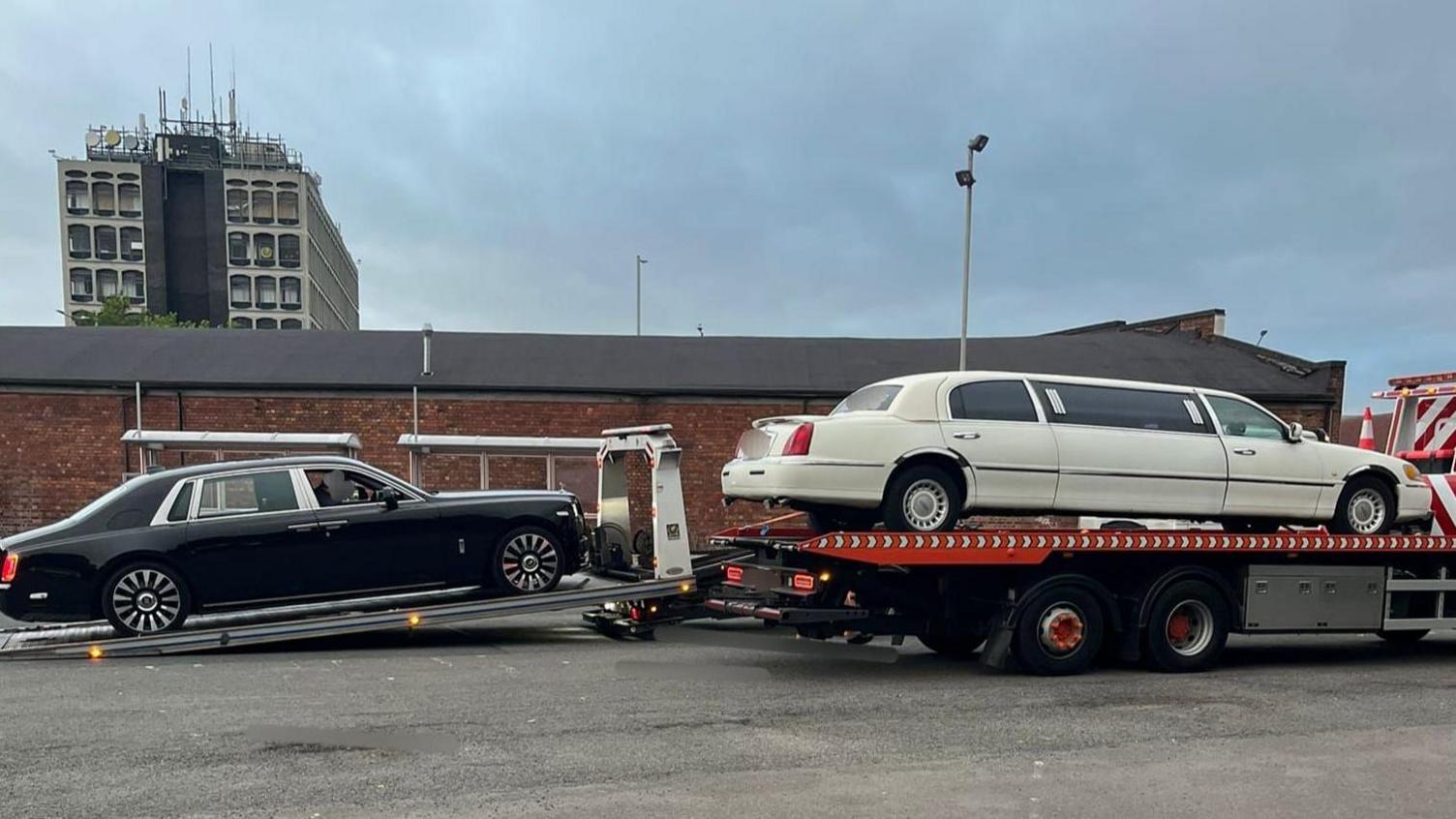 Two luxury cars on the back of a lorry. They are on a car park and made a lengthy convoy. There is little else in the picture apart from a brick building in the background and a grey tower block.