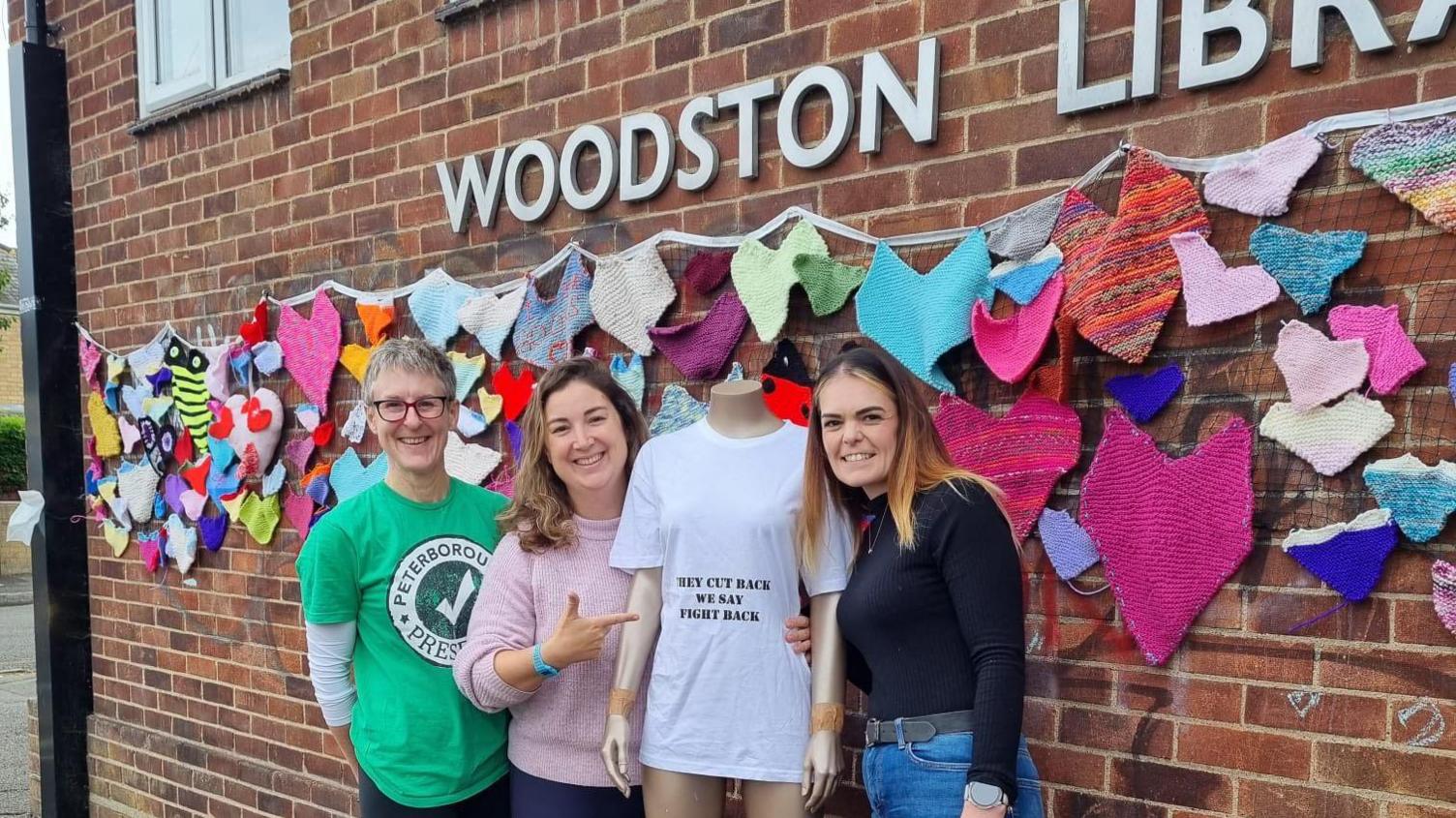 Volunteers at Peterborough Presents - a lady with a bob hair cut in a green T-shirt, a lady in a white jumper holding a mannequin wearing a white-T shirt standing next to another volunteer- a lady in a black jumper and blue jeans- all of them are standing in front a bricked wall with a Woodston Library signboard. 