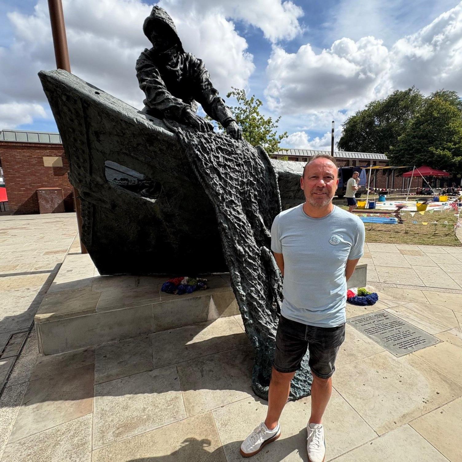 A man in his early 60s with short grey hair stands, wearing shorts and a t shirt, in front of a sculpture commemorating Grimsby's trawlermen. The sculpture comprises the bow of a boat, with a net spilling out.