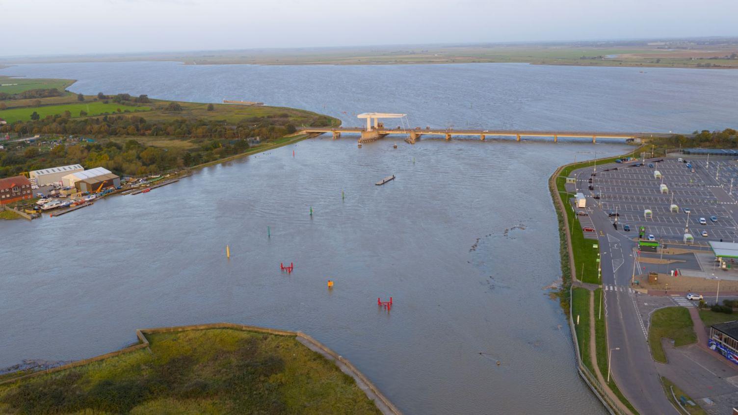 An aerial photo showing the vast expanse of the River Yare, with the large Asda supermarket car park to the right, and an industrial building to the left. The river flows under the A47 bridge.