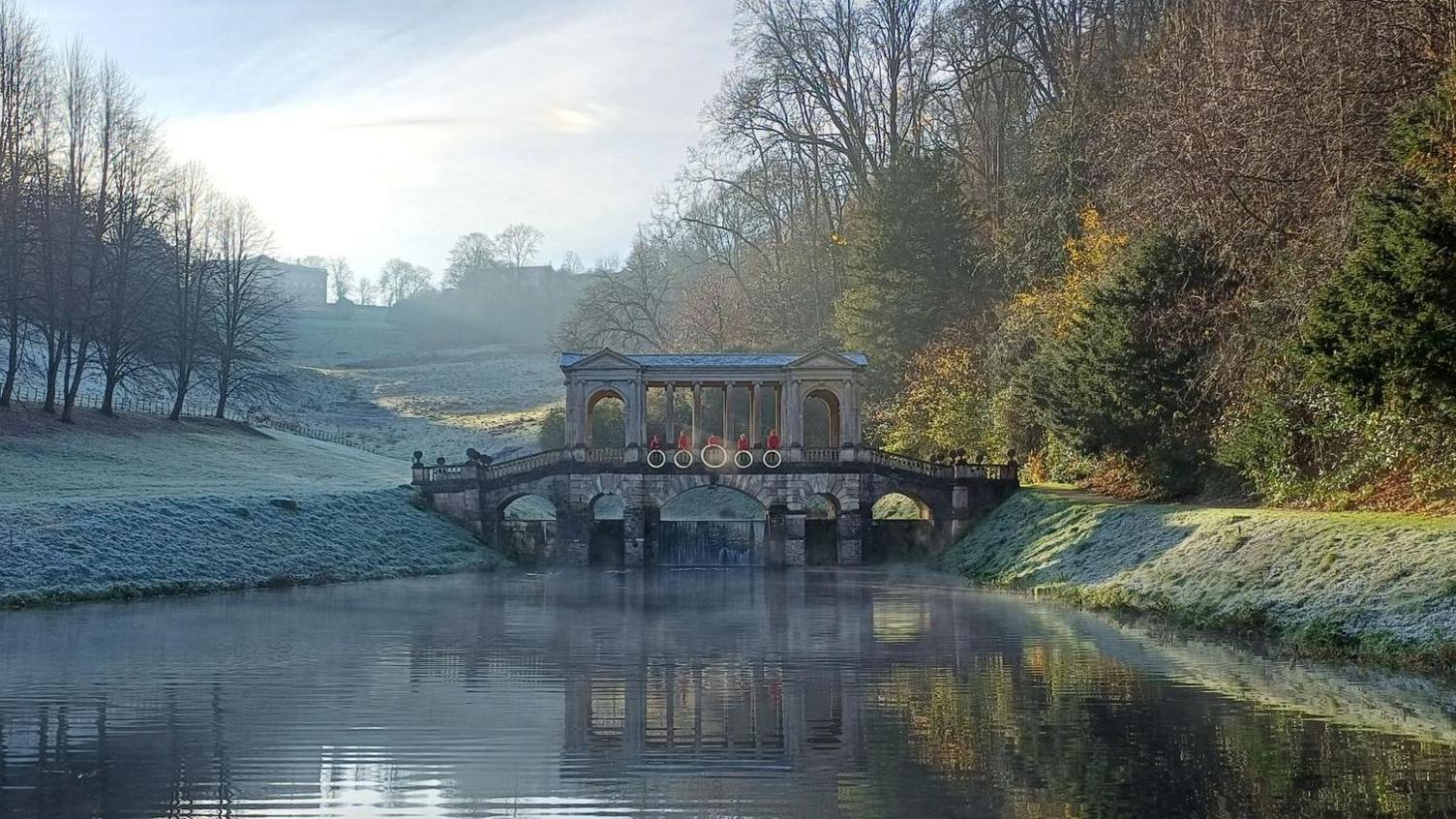 A tranquil image of a body of water surrounded by greenery with a small bridge over it. There are five people in red holding five golden hoops along the middle of the bridge.