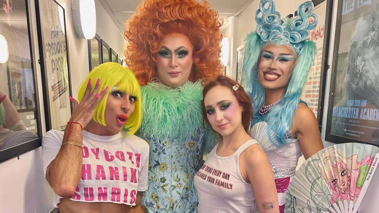 Shanika Sunrise wearing a yellow wig. They are standing next to two other drag queens, one with curly orange hair and the other with blue long hair.  The drag queens are standing with Chappell Roan who is wearing a white tank top and has red hair.