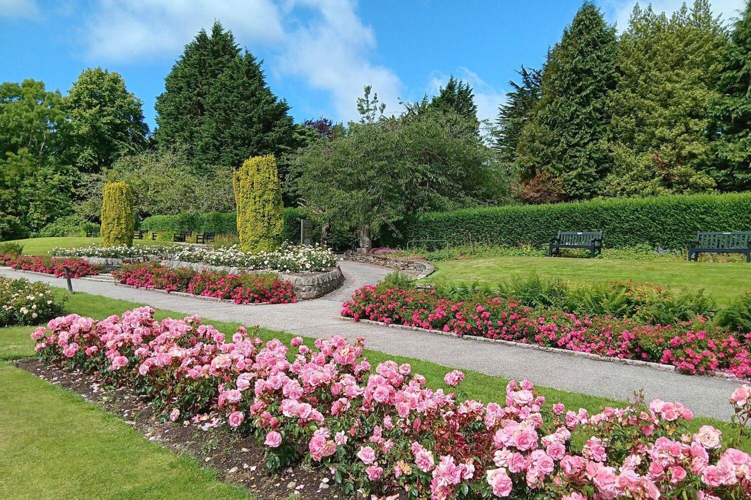 A manicured park with planted pink and red flowers. A gravel path runs around the neat flowerbeds and benches sit on the grass in the distance in front of a hedge.