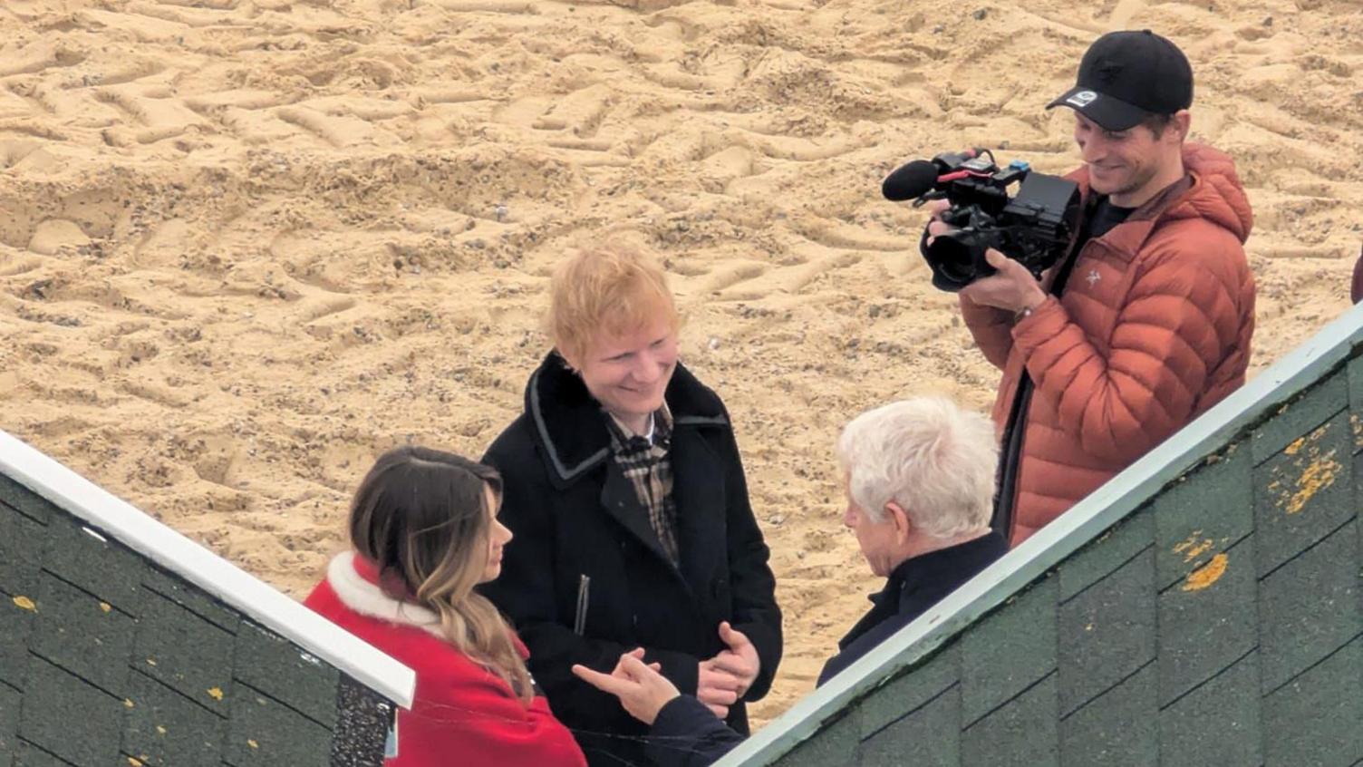 Ed Sheeran - a man with ginger hair wearing a black jacket - standing on a beach next to a woman wearing a red jacket and a man with a camera and a cap. They are talking to an elderly man with grey hair. 