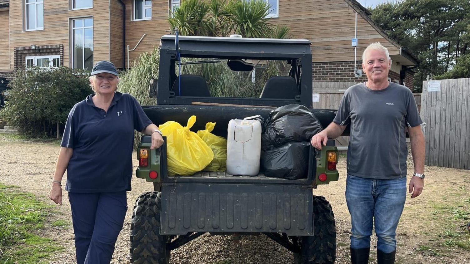 A woman in dark clothing and a cap stands to the left of a beach buggy parked on sandy ground, with the tailgate of the vehicle open and showing bagged waste which has been gathered, and man in a grey t-shirt and jeans stands to the right with a wooden cladded building in the background.