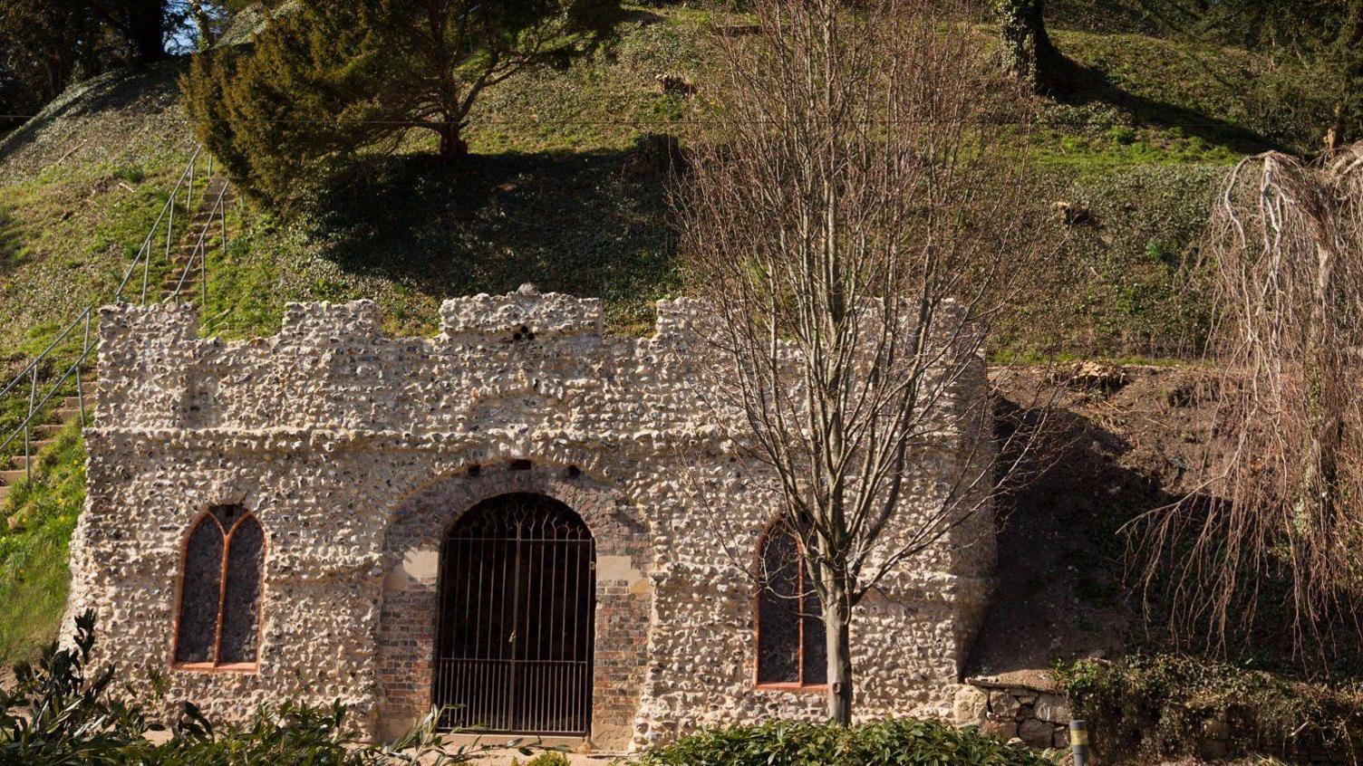 A small, castle-like structure made of small stones and brick with arched church-like windows and an arched gated entrance in the side of the mound.