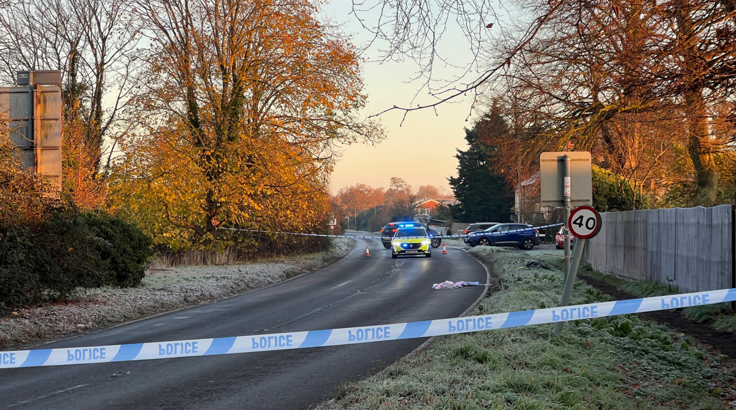 Police tape sections off part of a main road. Within the cordon looks to be fabric in the centre of the road, and another on the grassy verge at the side. 
Behind the cordon is a police car with flashing blue lights. 