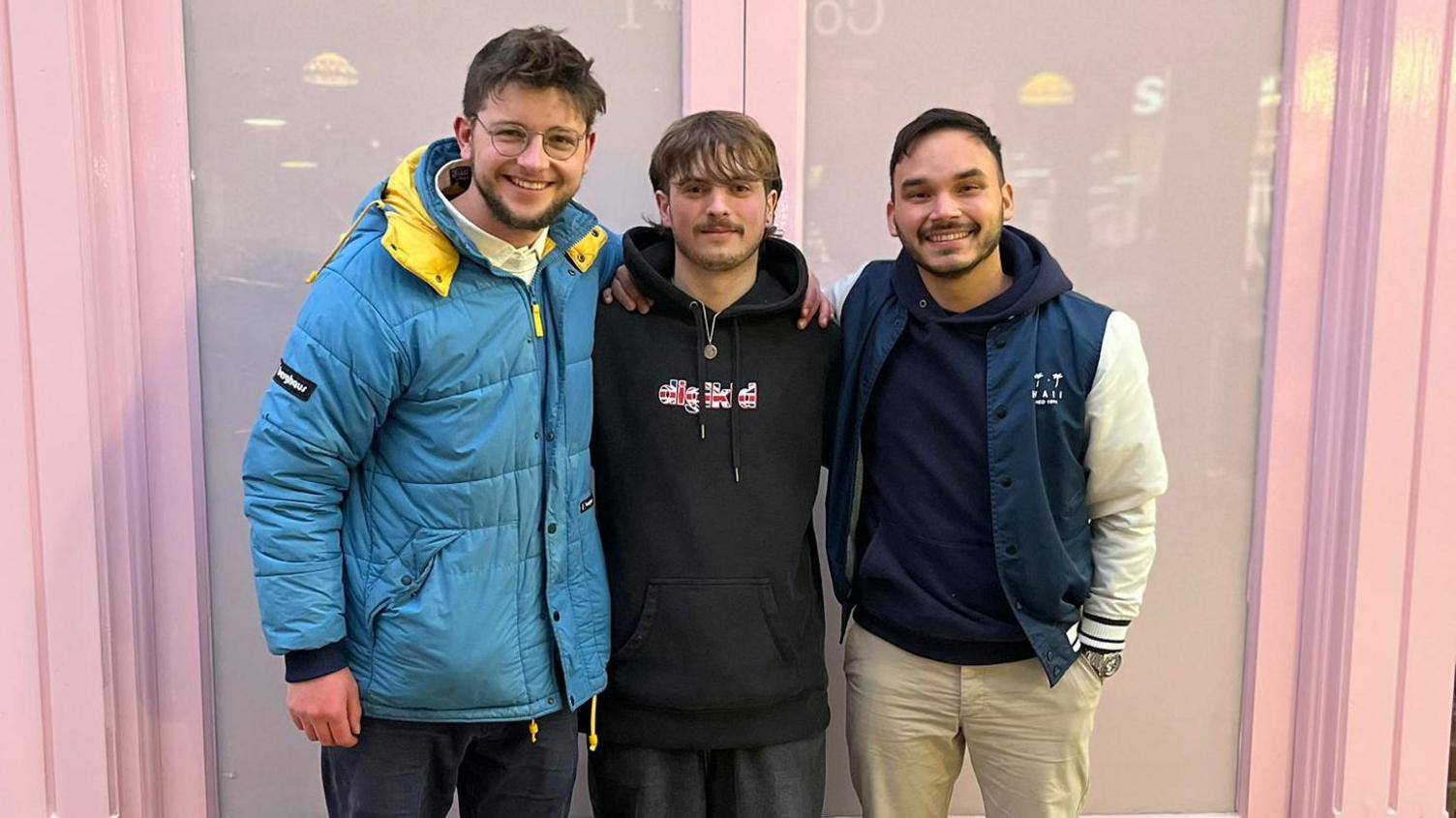 George Moir, Tathan and Isra stand with their arms around each other smiling at the camera. They're stood in front of a pair of pale pink double doors. George is on the left, he wears a pale blue hooded puffer jacket with has a bright yellow lining in the hood, a white collared polo shirt underneath and dark blue jeans. He has short spikey light brown hair with a short beard and wears think round wire framed glasses. Tathan stands in the middle, he has light brown hair which is styled into a fringe at the front with a moustahce and short beard. He wears a black hoodie which has some writing across the chest in a Union Jack print and charcoal grey tracksuit trousers. Isra is next to him on the right, he has short black hair and a short beard and wears a navy blue hoodie underneath a navy blue and white varisty style jacket and cream chinos.