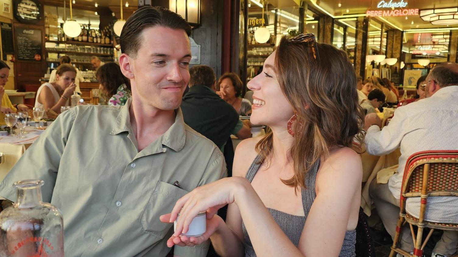 Alasdair and Eloïse looking at each other and smiling in a restaurant. He is wearing a short sleeved green shirt and she has a strappy grey top on with large earrings and sunglasses on her head. 